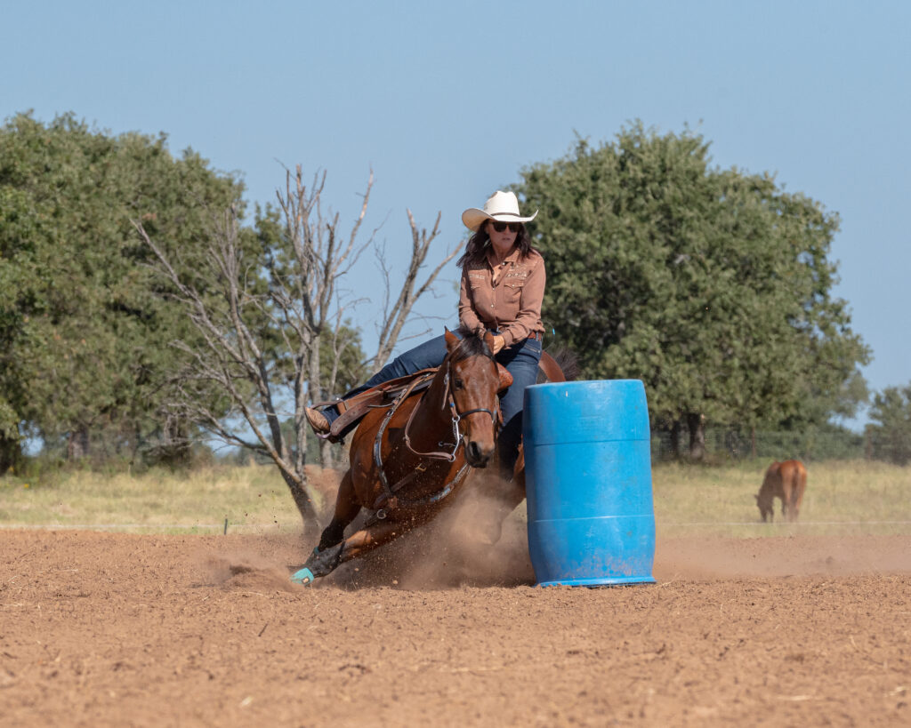 Barrel racer in a white cowboy hat rounds a blue barrel on a sorrel horse, kicking up dirt in an outdoor arena