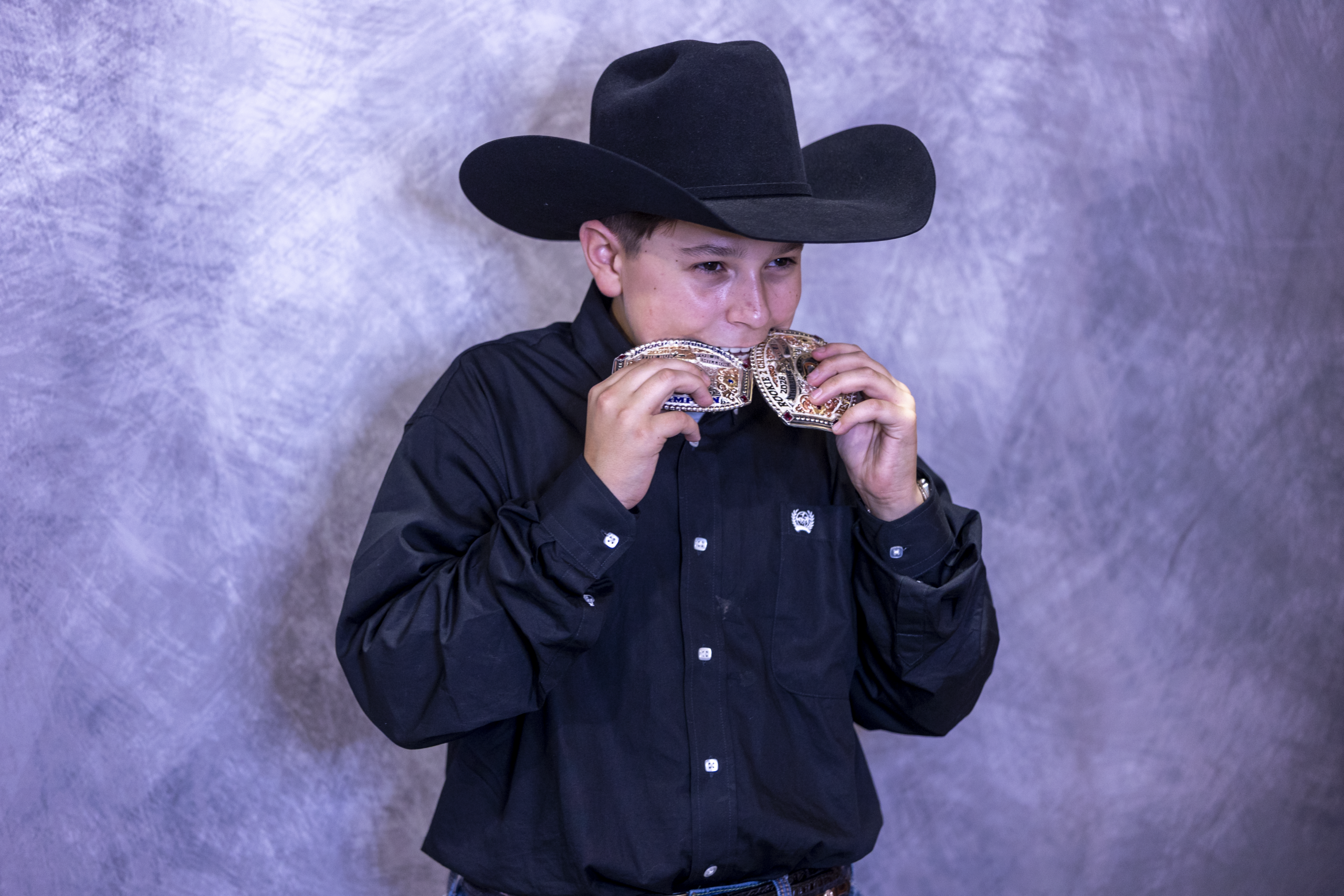 Rodeo competitor in black western shirt and cowboy hat holds multiple gold medals around his neck against a purple background