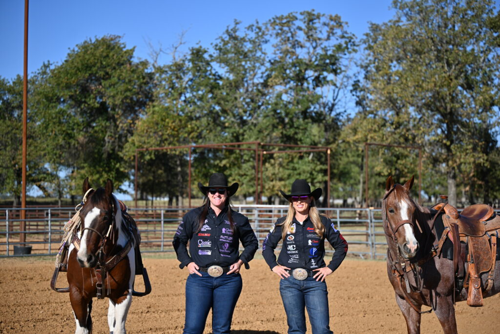 Two women in black cowboy hats and sponsor-patched shirts stand hands on hips in a dirt arena between two saddled horses