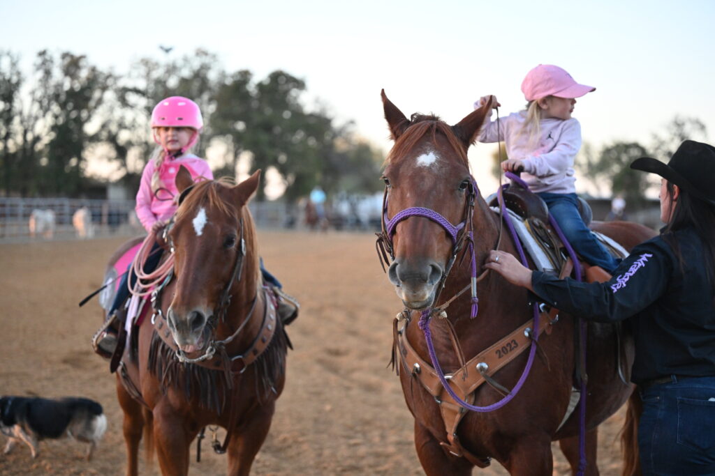 Two young riders in pink helmet and ball cap sit aboard sorrel horses as a woman in a cowboy hat leads one at dusk