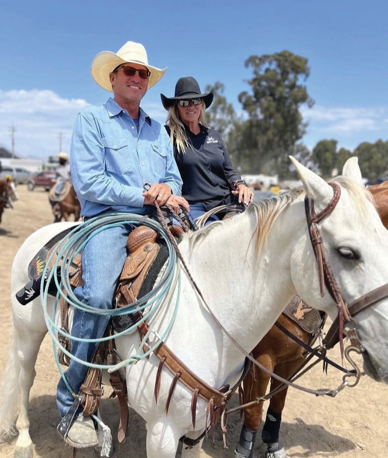 Man in straw cowboy hat on a gray horse with a coiled rope beside a woman in a black hat on a sorrel at a rodeo