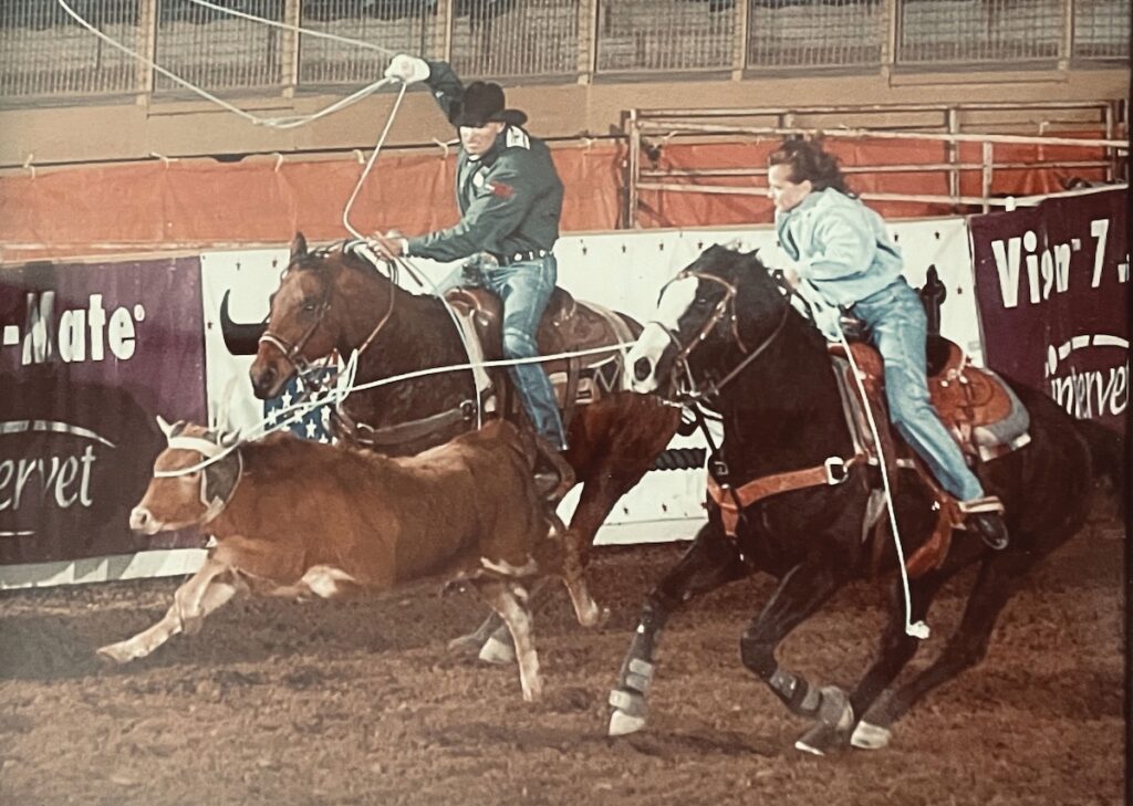 Team roping run in progress as a header on a sorrel horse swings his loop and a heeler on a dark horse chases the steer