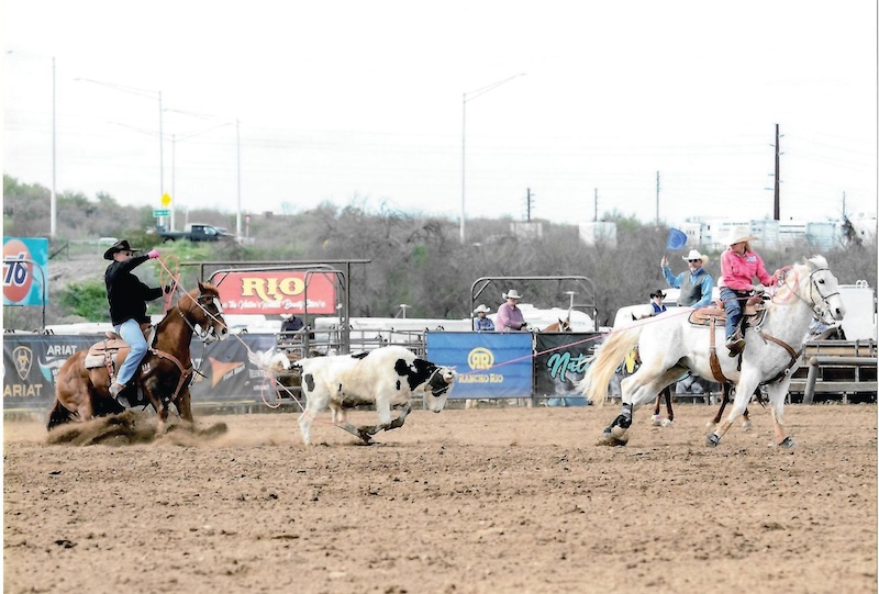 Team ropers on sorrel and gray horses work a steer in a dirt arena in front of Rio sponsor banners