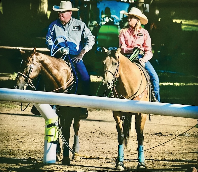 Two riders in cowboy hats sit horseback behind an arena rail, the man coiling a rope while a woman in a pink shirt looks off to the side