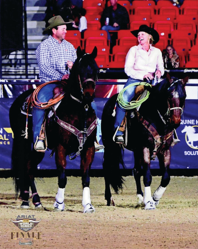 Two team ropers in cowboy hats share a laugh on horseback in the arena, coiled ropes hanging from their saddles