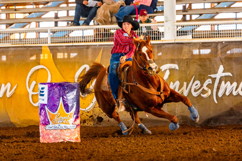 Barrel racer in a red striped shirt and black cowboy hat rounds a Royal Crown barrel on a sorrel horse in an indoor arena