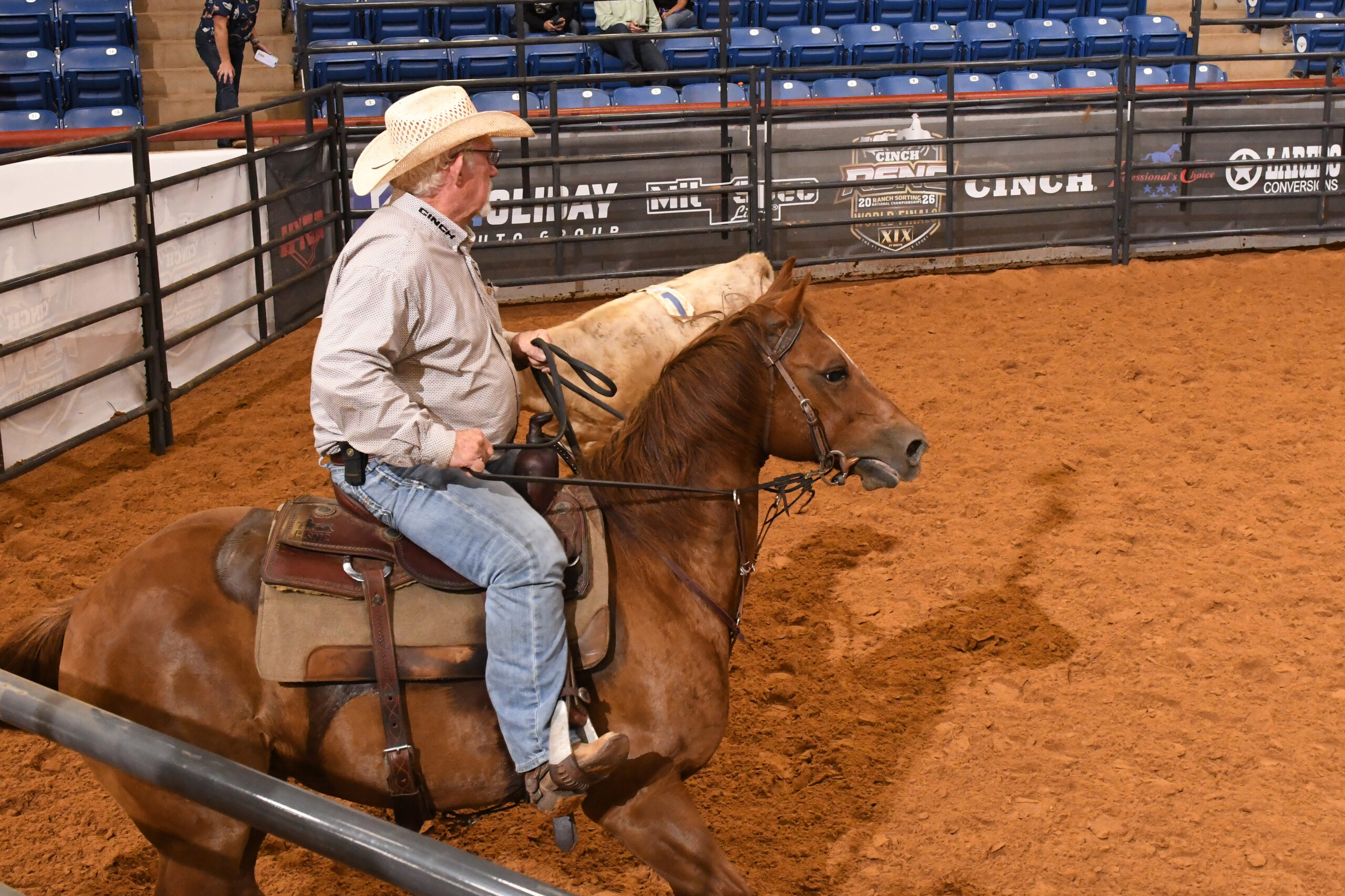 Cowboy on saddled brown horse in indoor rodeo arena with spectator seating visible behind metal fencing