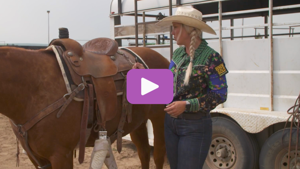 Blonde cowgirl in a straw hat and floral western shirt standing beside a saddled sorrel horse near a stock trailer