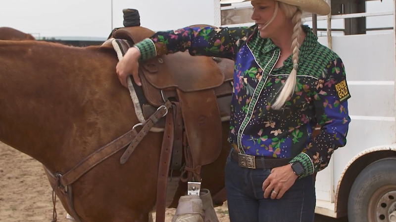 Blonde cowgirl in a floral western shirt and straw hat rests her hand on a saddled sorrel horse beside a trailer