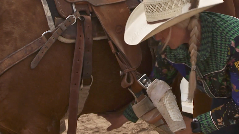 Cowgirl in a straw hat and braid bends down beside her sorrel horse's saddle to adjust a stirrup and boot