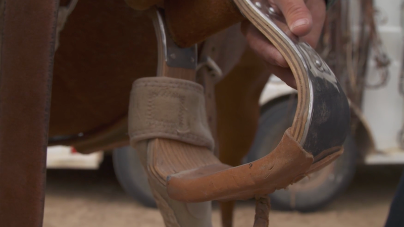 Close-up of a hand gripping a wide leather-wrapped stirrup on a western saddle, with a horse's leg behind