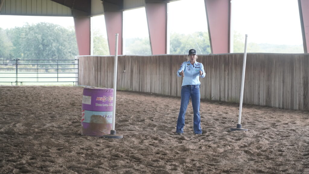 Woman in a light blue western shirt and ball cap speaks in a covered arena beside a barrel and training poles