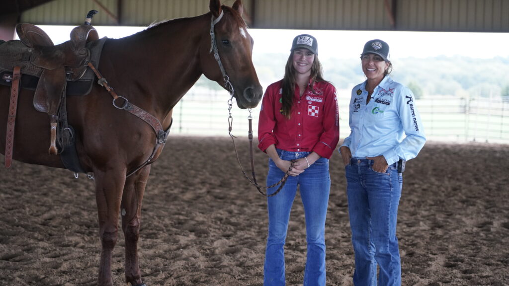 Two smiling women in ball caps and sponsor shirts stand beside a saddled sorrel horse in a covered arena