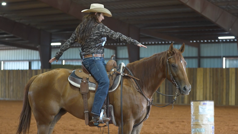 Rider in a straw cowboy hat and camo shirt stretches both arms out wide while mounted on a sorrel horse in an indoor arena