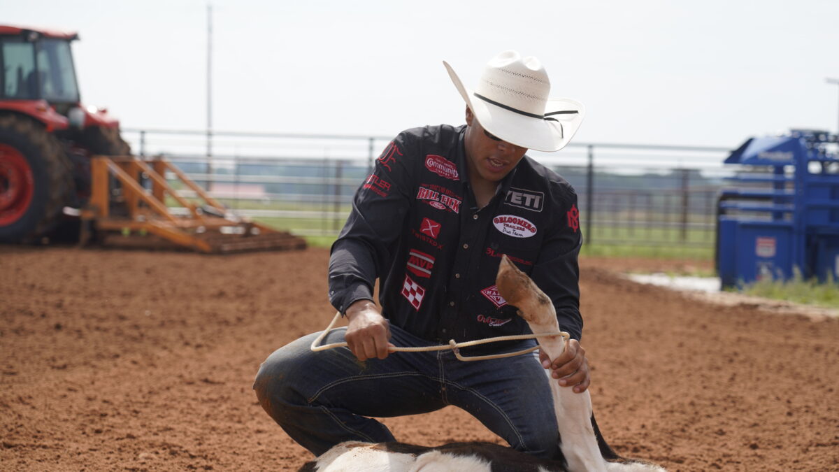 Cowboy in a white straw hat and sponsor-covered black shirt kneels in the dirt arena to tie a calf with a piggin' string