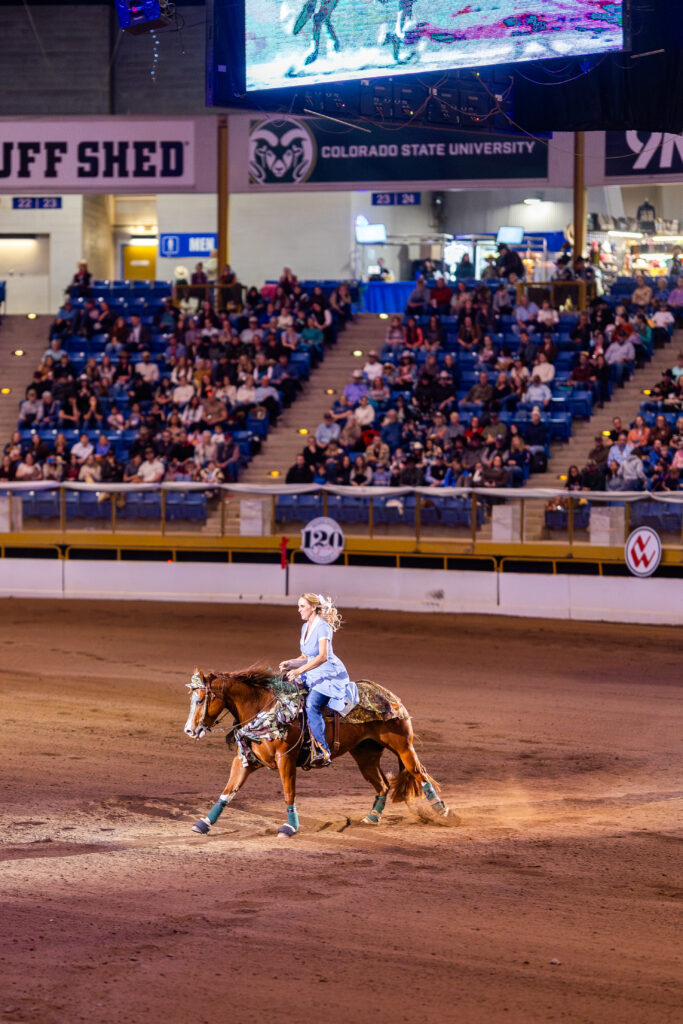 Rider in a blue dress gallops a chestnut horse across the arena dirt as crowds watch from Colorado State University stands