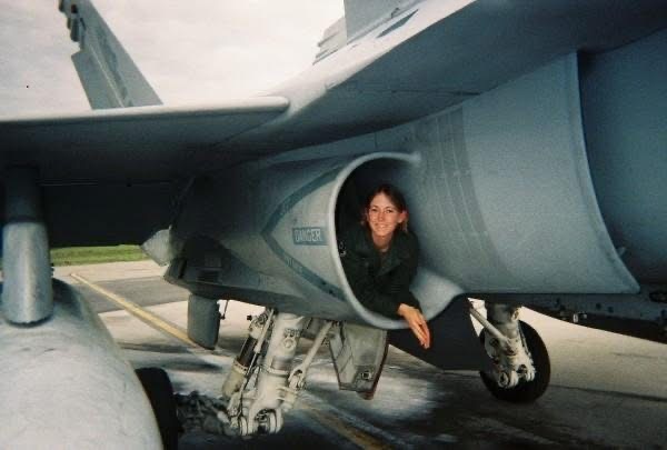 Smiling woman peeking out from inside the jet intake of a fighter aircraft parked on a tarmac