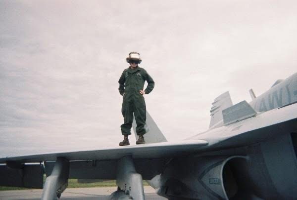 Person in a flight suit and helmet stands with hands on hips atop the wing of a fighter jet under gray skies