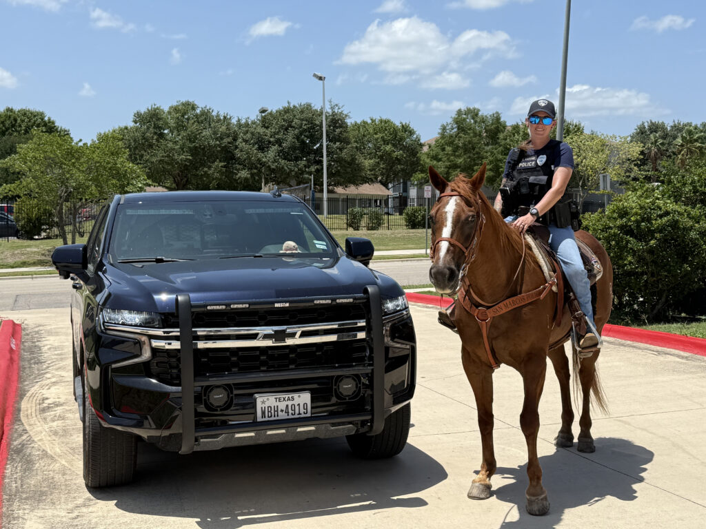 Mounted police officer in sunglasses and vest rides a sorrel horse alongside a black patrol SUV on pavement