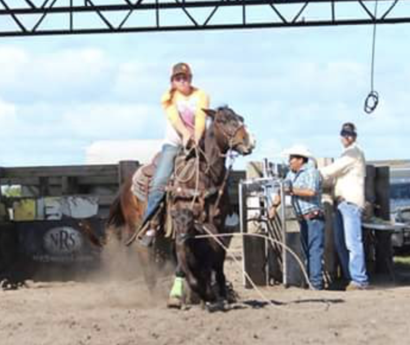 Young rider in a yellow shirt and cap stays seated as her bay horse bucks in a dusty arena while two cowboys watch from the chute