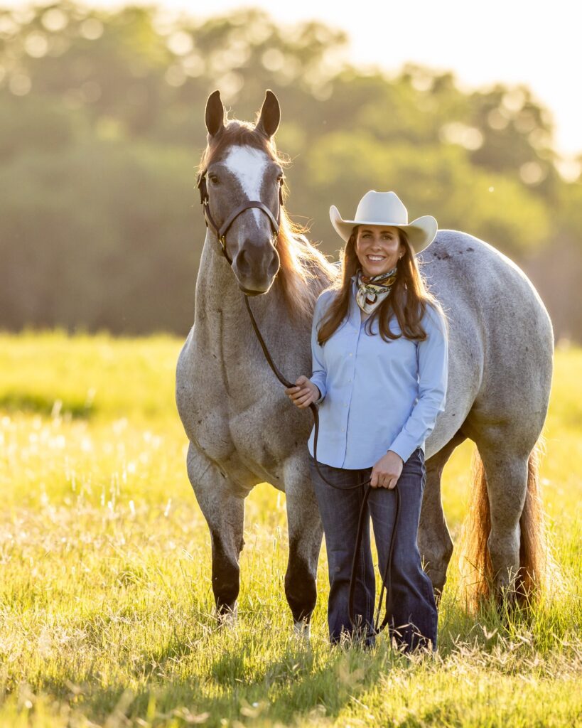 Woman in cowboy hat and western attire stands with gray horse in sunny pasture