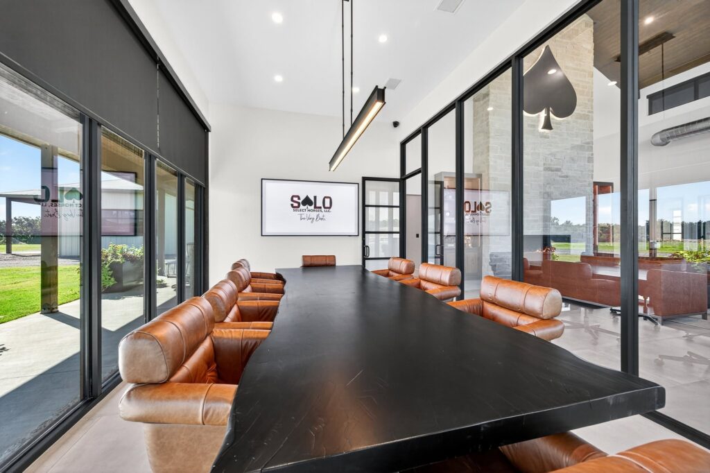 Modern equestrian facility interior with "SOLO" signage, featuring a black conference table surrounded by cognac leather seating and floor-to-ceiling windows overlooking pastures