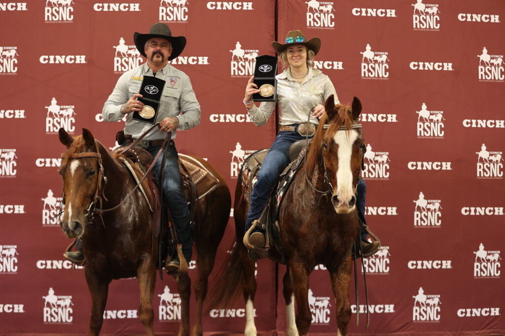 Two riders on horseback hold up champion buckles in presentation cases in front of a Cinch RSNC backdrop