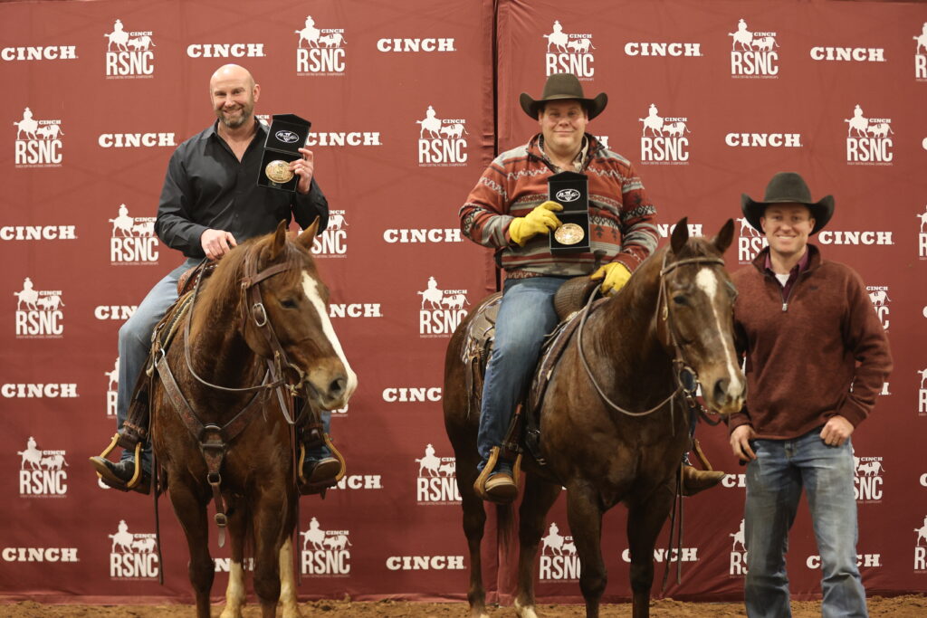 Two mounted riders hold up championship buckles in presentation cases beside a man standing at right, in front of a Cinch RSNC backdrop