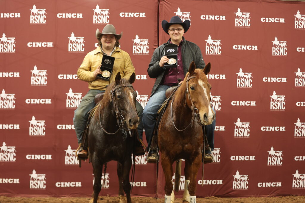 Two mounted riders in cowboy hats hold up belt buckle award cases in front of a Cinch RSNC backdrop