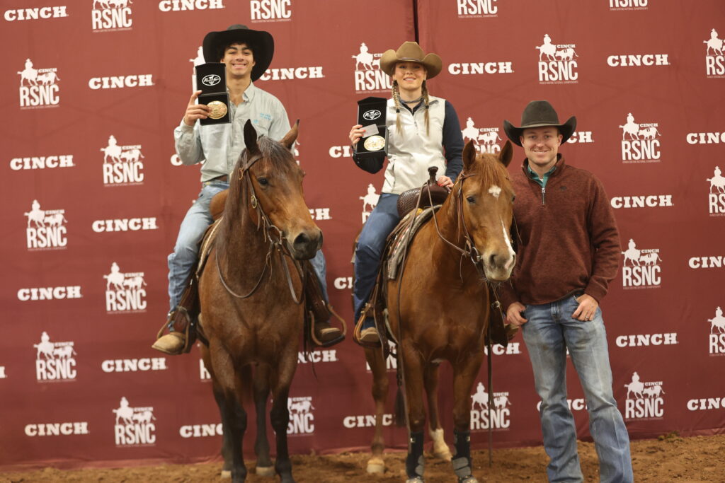 Two mounted riders hold up buckle awards beside a man standing on the ground in front of a Cinch RSNC backdrop