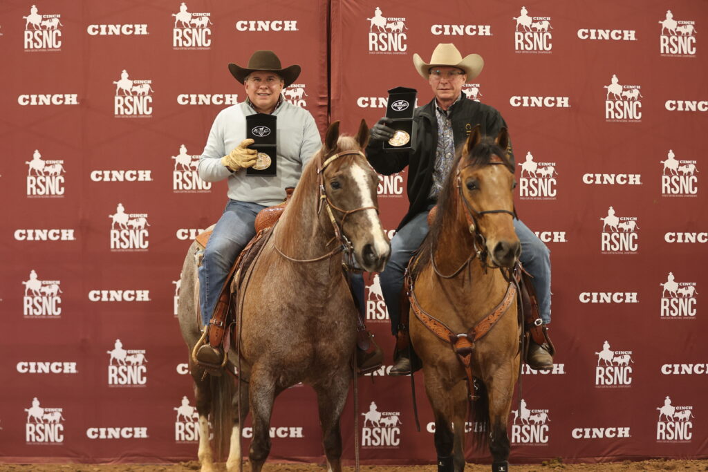 Two mounted riders in cowboy hats hold up award buckles in front of a Cinch RSNC backdrop