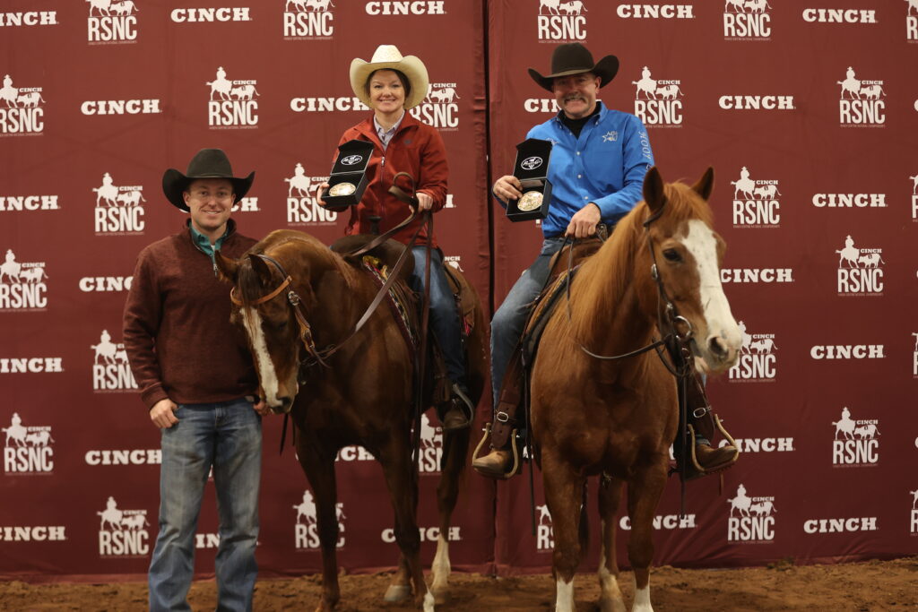 Two mounted riders hold up belt buckle award cases beside a man standing at a horse's head in front of a Cinch RSNC backdrop