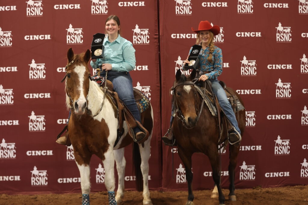 Two young riders hold up championship buckle cases on horseback in front of a Cinch RSNC backdrop
