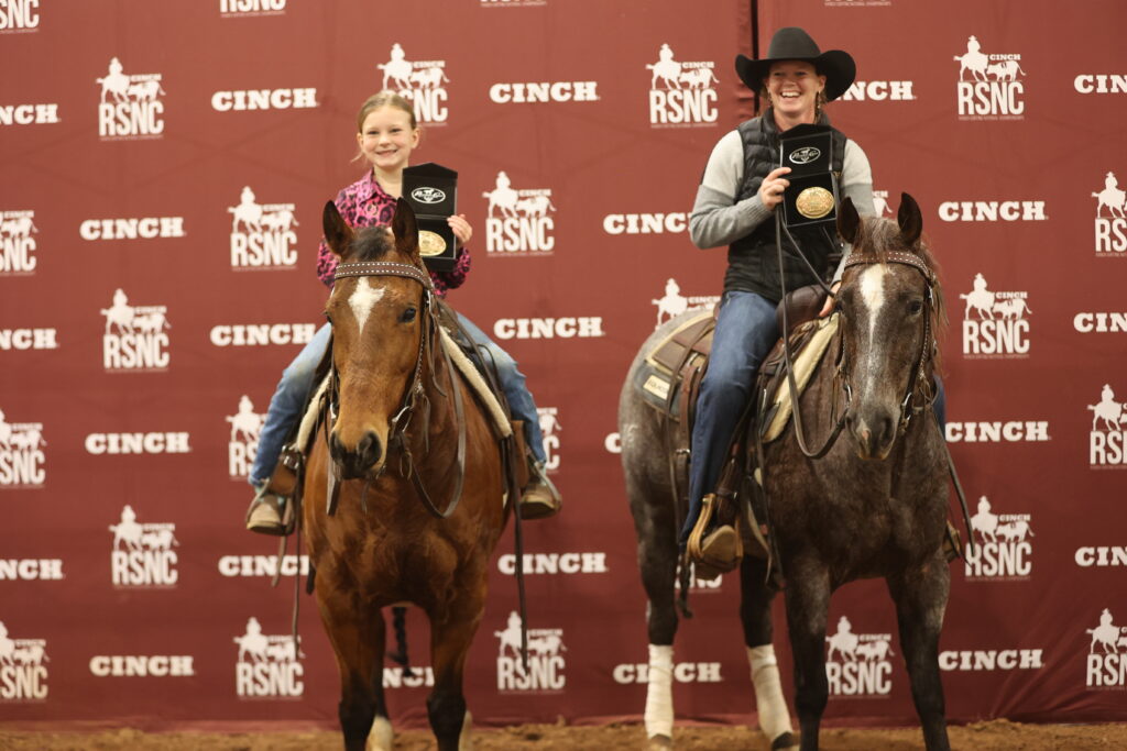 Two riders on horseback hold up championship buckles in front of a Cinch RSNC backdrop