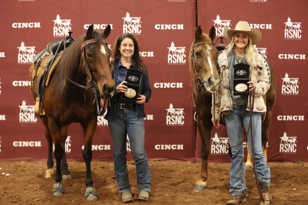 Two women hold championship belt buckles beside their saddled horses in front of a Cinch RSNC backdrop