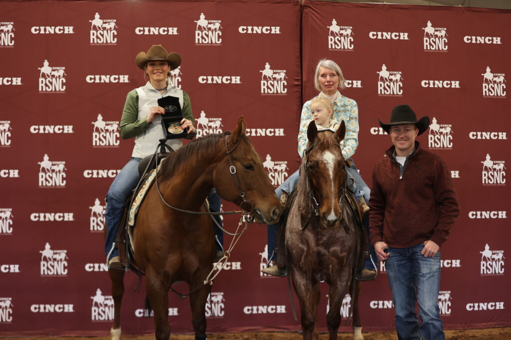 Two mounted riders, one holding a trophy buckle and one with a child in the saddle, pose beside a man on foot in front of a Cinch RSNC backdrop