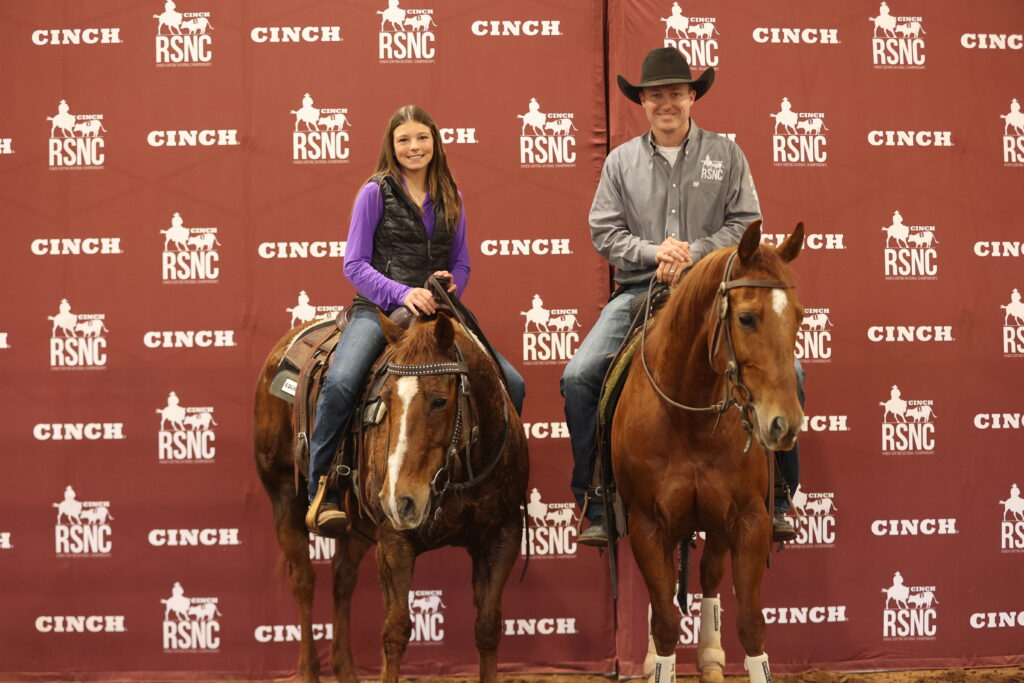 Young rider in a purple shirt and vest and a man in a cowboy hat sit mounted on sorrel horses in front of a Cinch RSNC backdrop