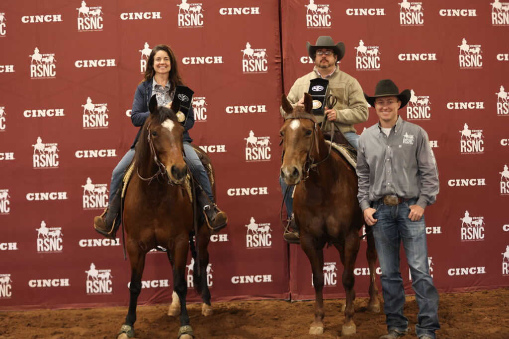 Two mounted riders hold buckle presentation cases beside a standing man in front of a Cinch RSNC backdrop