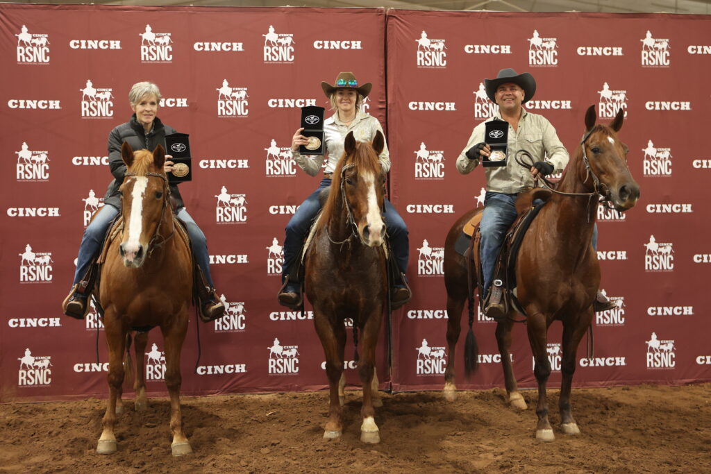 Three riders on horseback hold up belt buckle award cases in front of a Cinch RSNC backdrop in an indoor arena