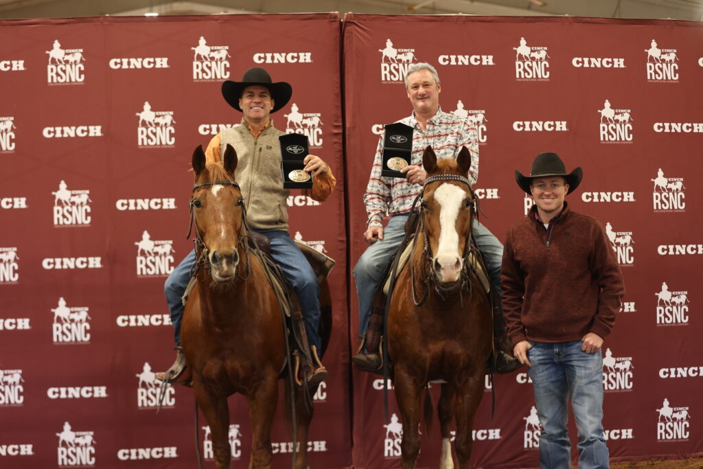 Two riders on horseback hold championship belt buckles beside a standing man in front of a Cinch RSNC backdrop