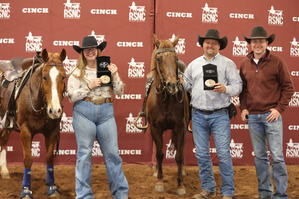 Two winners hold trophy buckles beside their sorrel horses as a third man stands by, in front of a Cinch RSNC backdrop