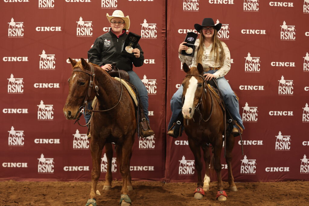Two mounted riders in cowboy hats hold up award buckles in front of a Cinch RSNC backdrop