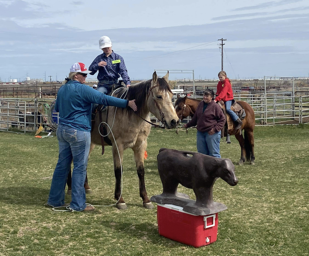 Instructor in a red cap steadies a buckskin horse as a helmeted young rider practices roping a dummy steer on a cooler