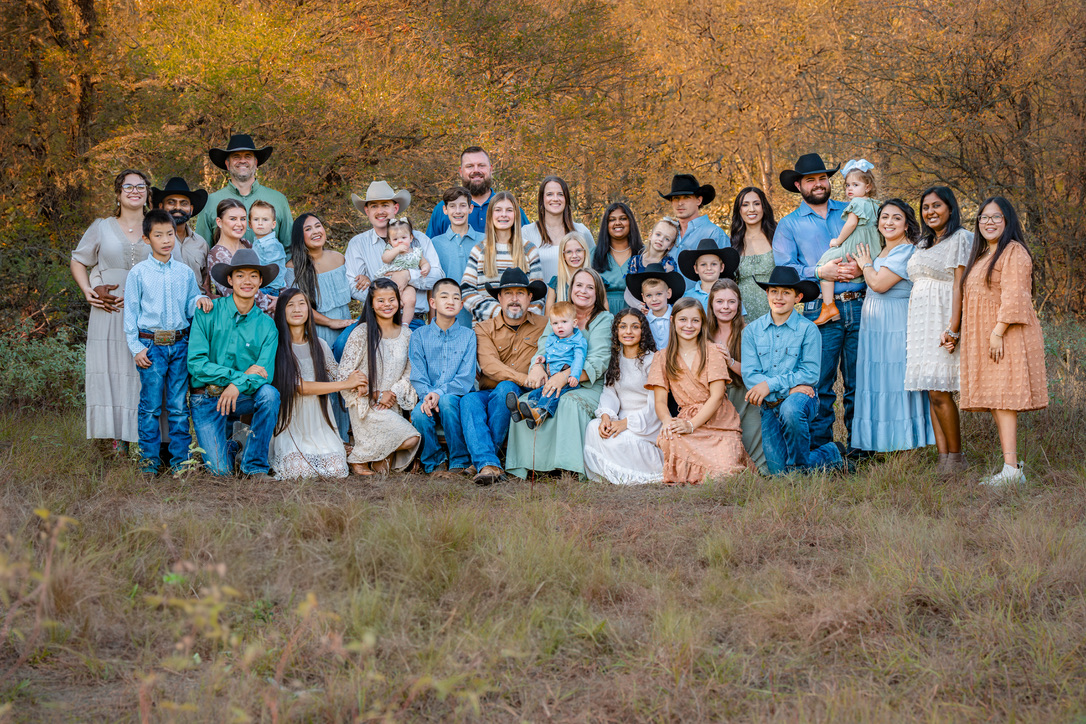 Large extended family wearing coordinated blue and neutral tones poses together outdoors against a brown textured backdrop