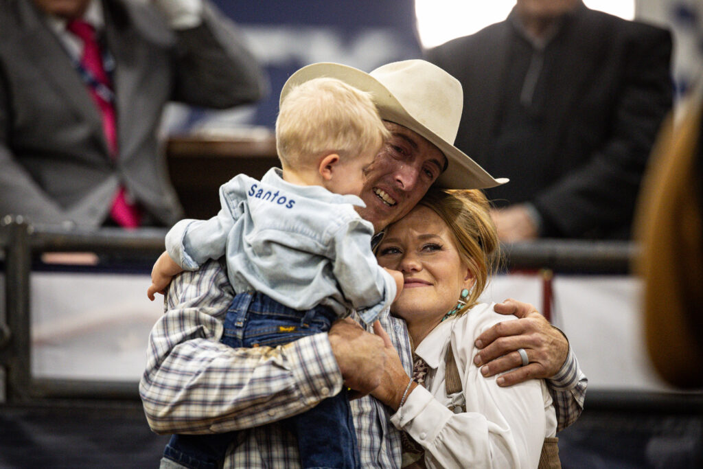 Cowboy in white hat holds young child wearing "Santos" shirt while standing with woman in western attire at indoor rodeo arena