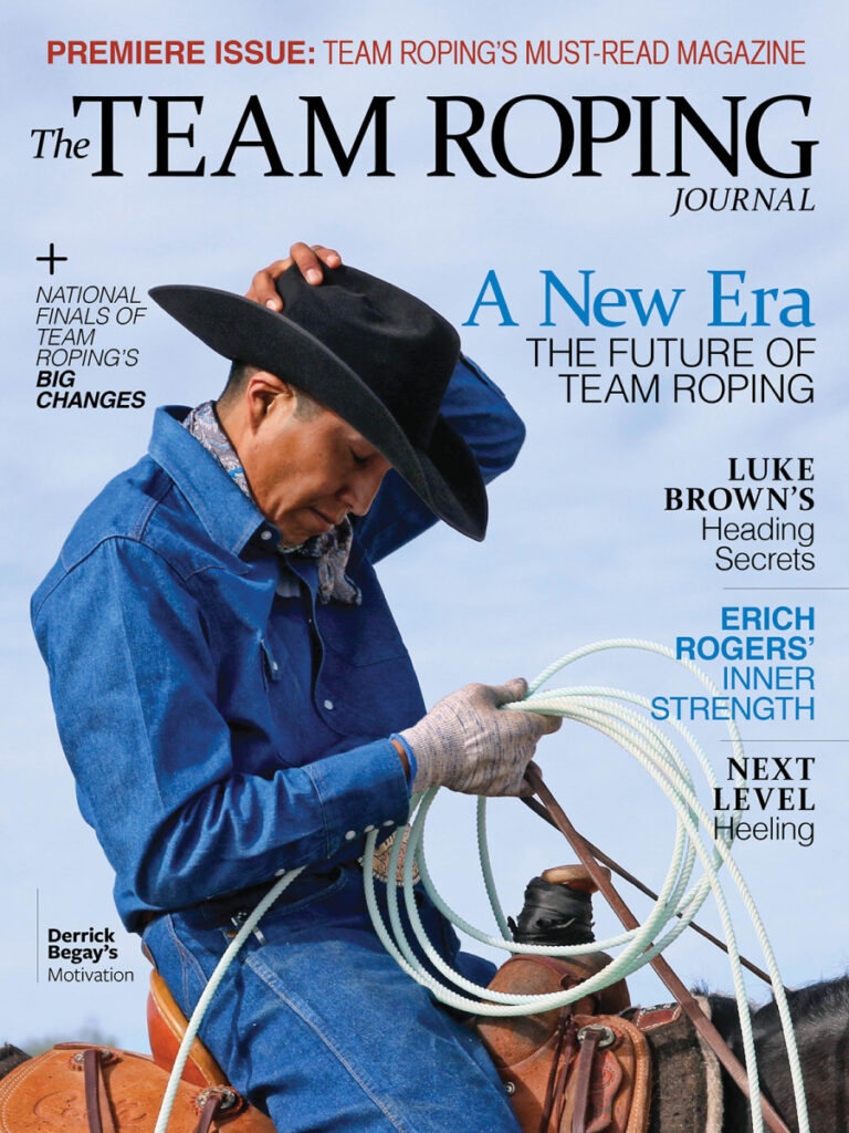 Cowboy in blue shirt and black hat holds a coiled rope while standing next to a saddle, on the cover of The Team Roping Journal premiere issue