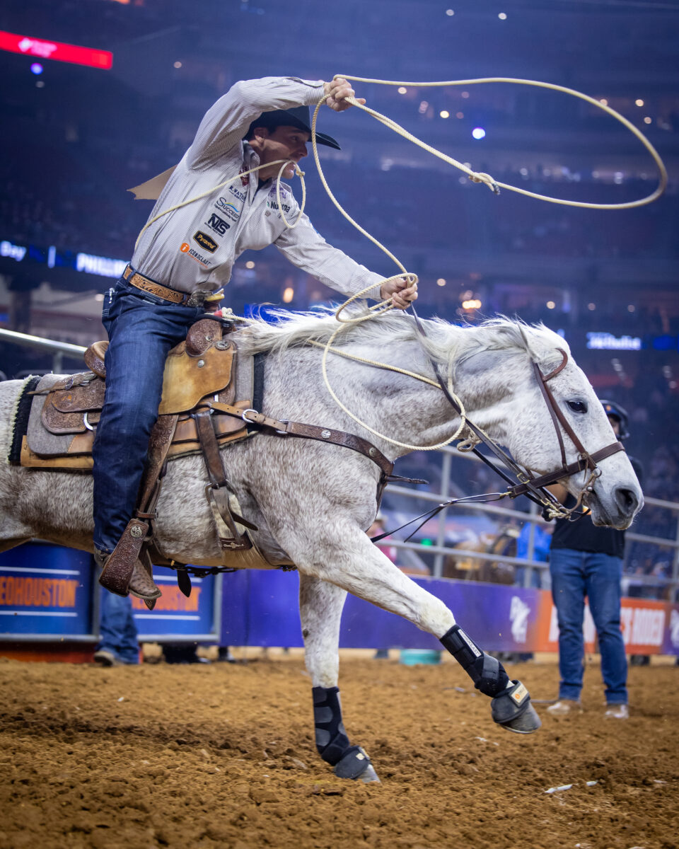 Cowboy in a black hat swings his loop overhead aboard a gray horse in a dirt arena during a rodeo roping run