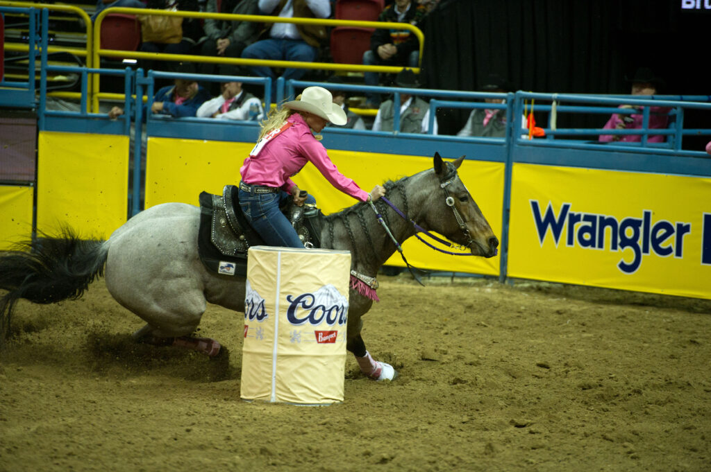 Barrel racer in a pink shirt and white cowboy hat turns a roan horse tightly around a Coors barrel in an indoor arena