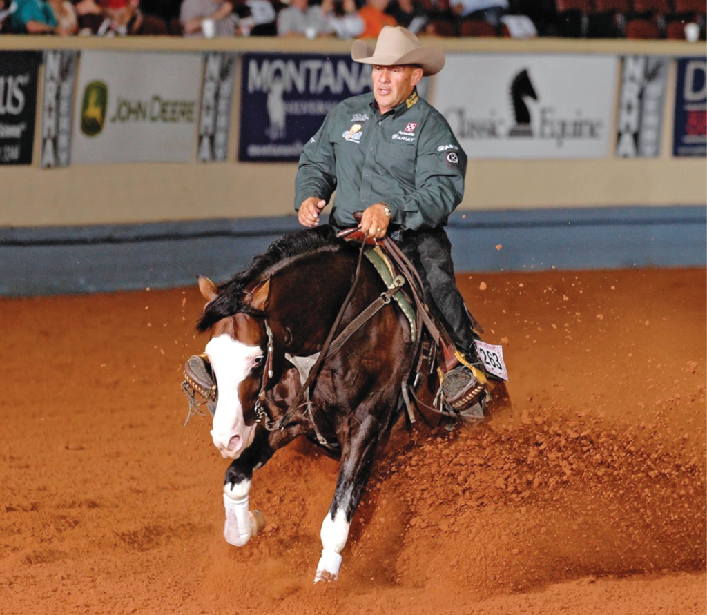 Cowboy in a straw hat and green shirt sliding his bay reining horse to a dramatic stop, spraying red arena dirt
