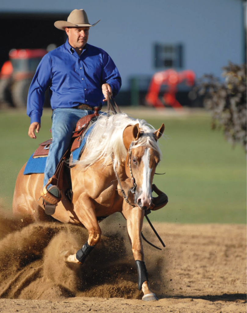 Cowboy in a blue shirt and straw hat sliding to a stop on a palomino reining horse in a cloud of dirt